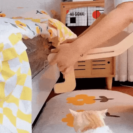 Hands installing a window hammock for cat with a wooden frame while a ginger cat watches below